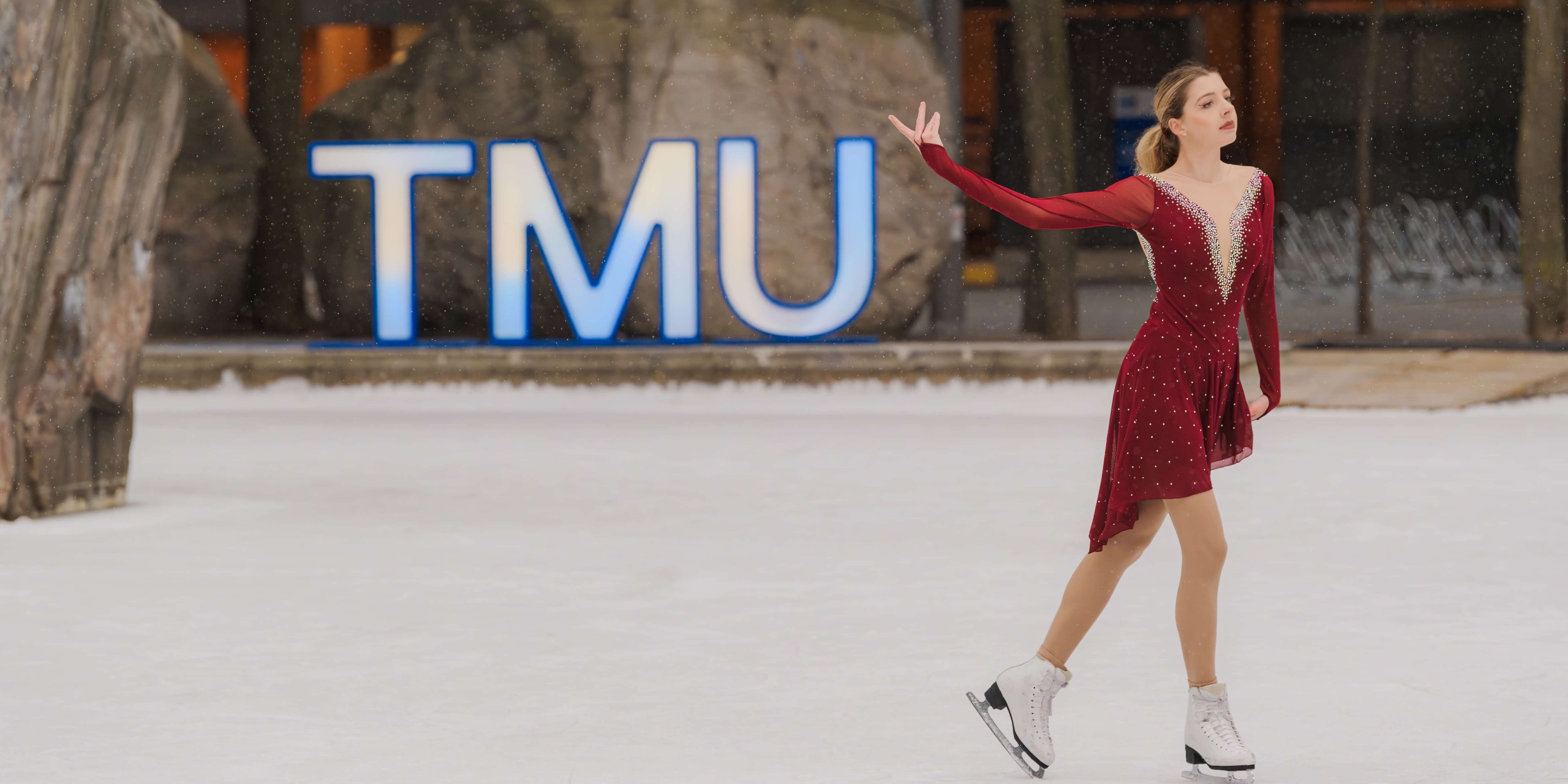 A figure skater wearing a red dress performs on Lake Devo ice rink. The TMU logo light up sign in the background.